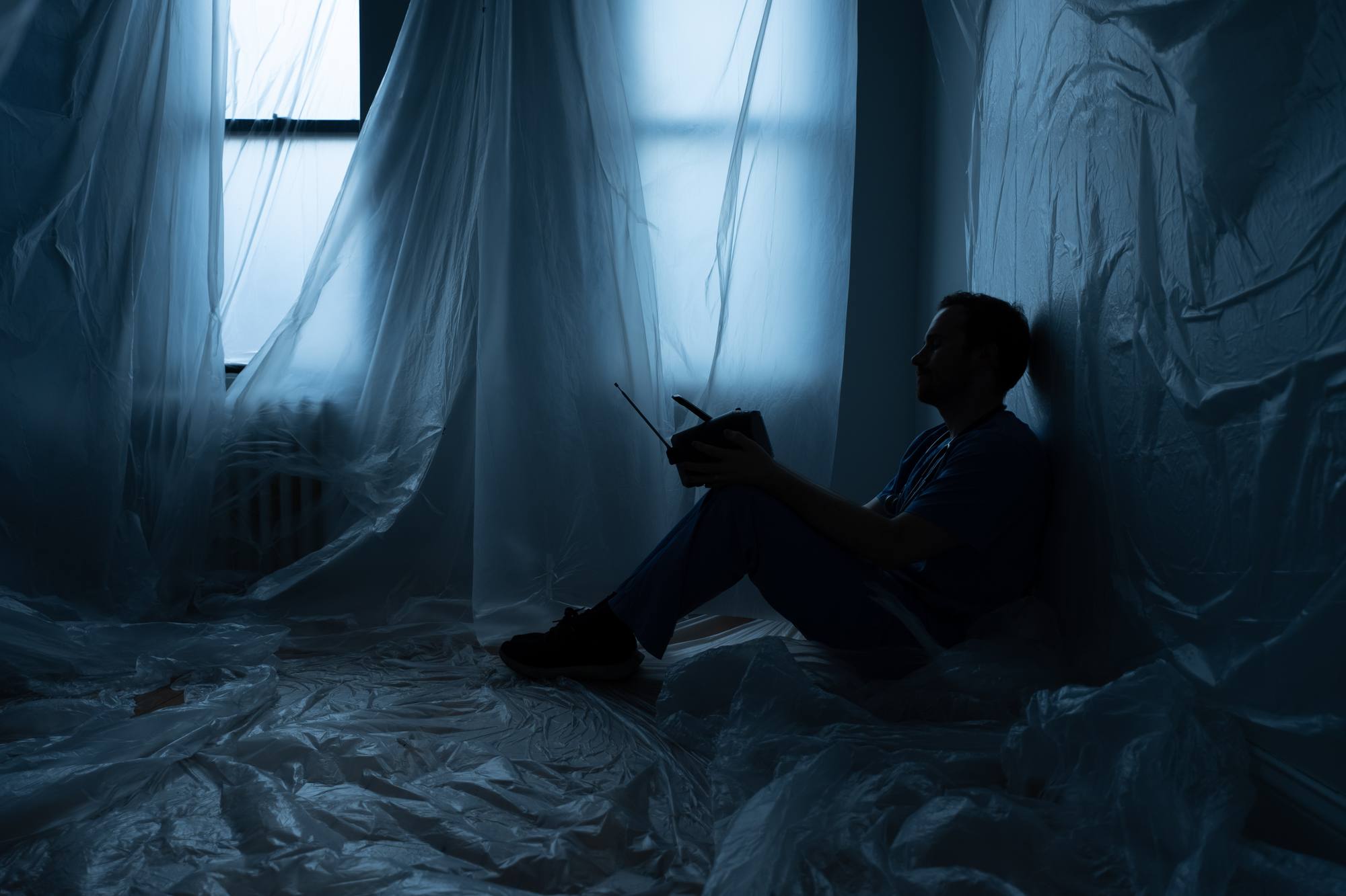 J Lind in scrubs, sitting on the floor against a wall in a dim blue room draped in plastic sheeting, holding a small radio