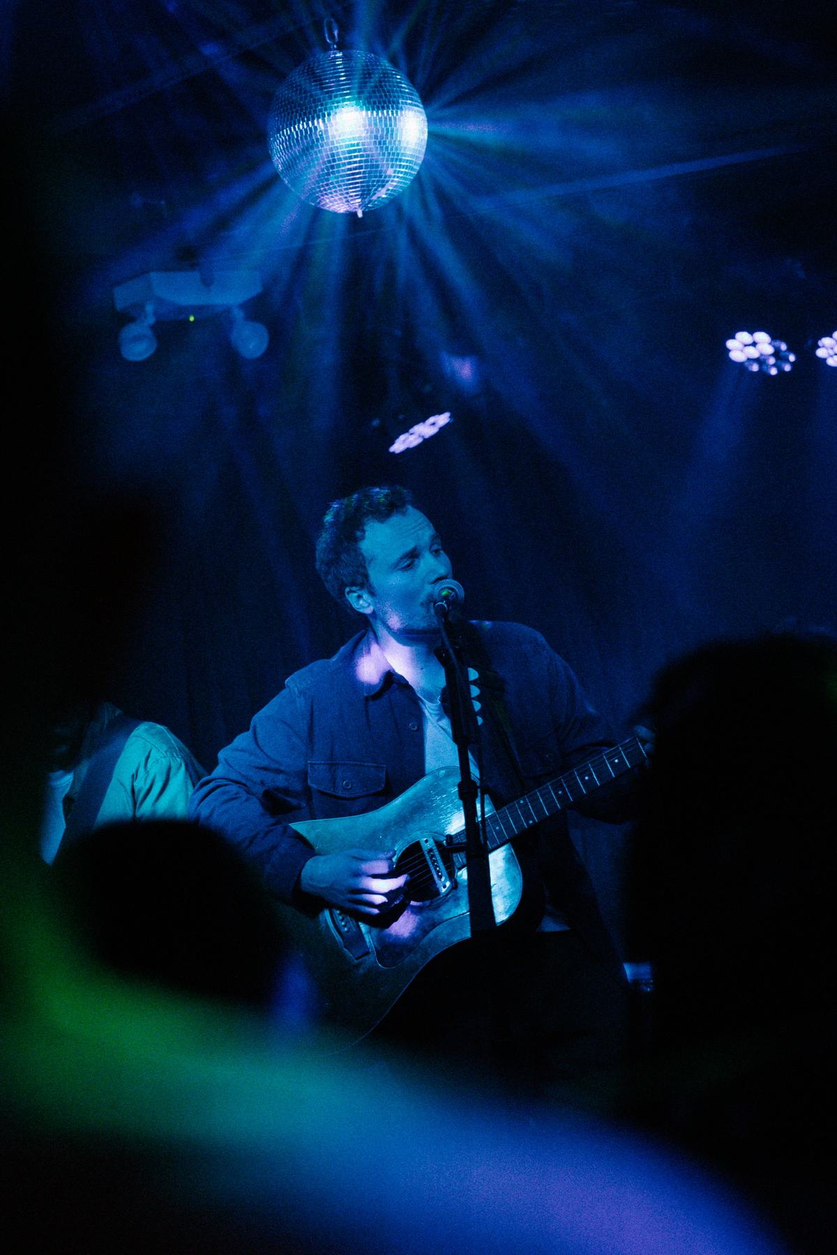J Lind singing and playing acoustic guitar on stage under a mirror ball, lit in blue