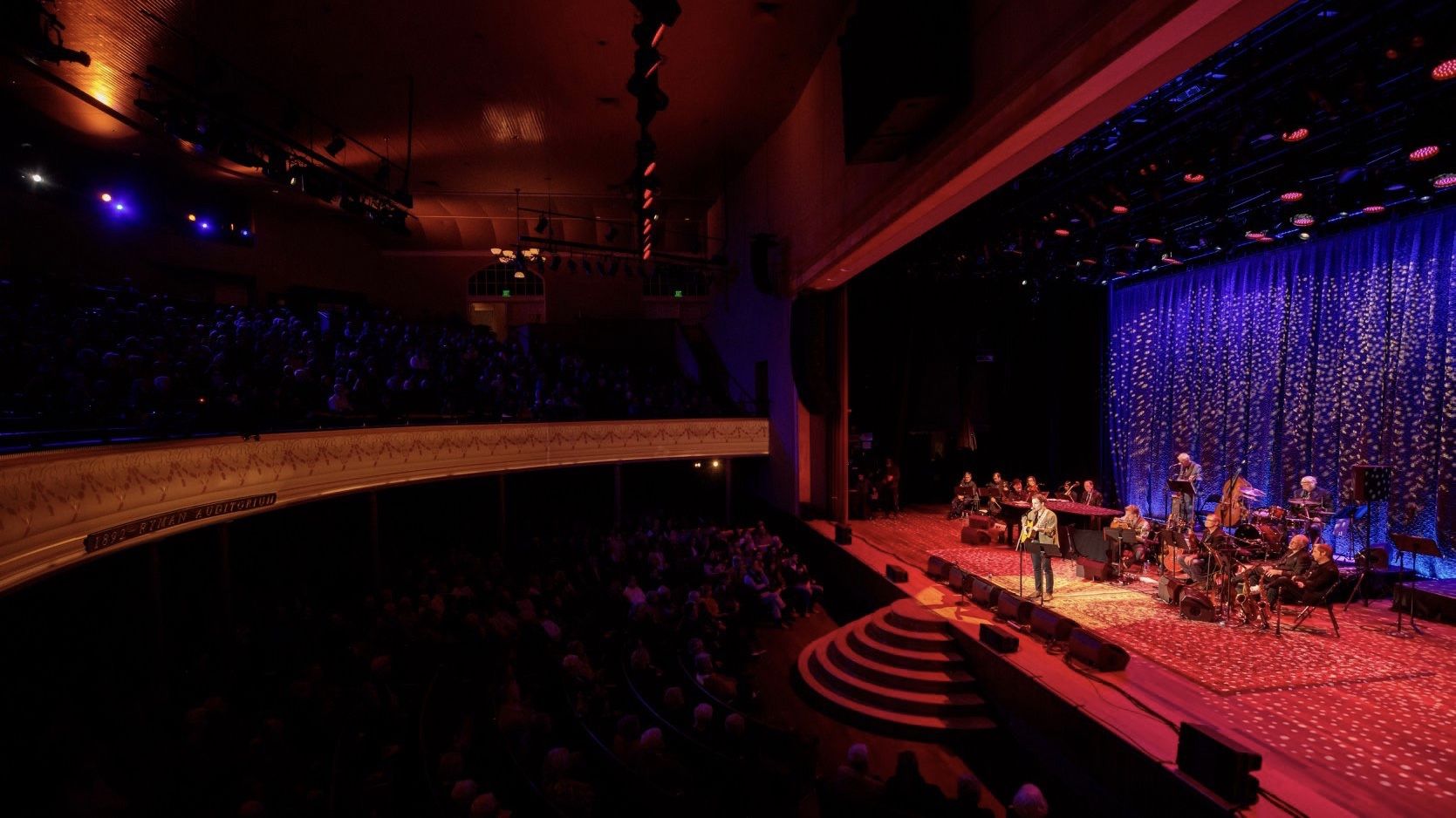 J Lind at the microphone with his guitar on the Ryman Auditorium stage, a band behind him and a full house in the dark